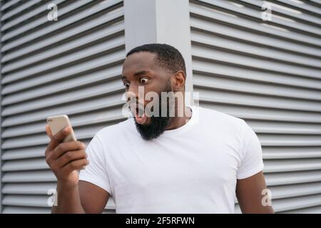 Choqué l'homme américain africain avec la bouche ouverte en utilisant le téléphone mobile regardant l'écran numérique. Un gars stupéfait de regarder des nouvelles en ligne Banque D'Images