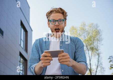 Jeune homme enthousiaste utilisant un smartphone, jouant à un jeu mobile. Portrait du gagnant de loterie en ligne surjoyeuse regardant l'appareil photo. Concept de Paris sportifs Banque D'Images