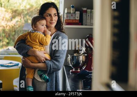 Mère et enfant ensemble dans la cuisine donnant sur la fenêtre extérieure. Mère transportant son bébé debout à la maison. Banque D'Images