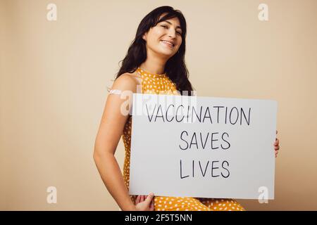 Belle femme tenant un panneau avec "la vaccination sauve des vies" écrit dessus. Femme avec bandage sur son bras après avoir reçu la vaccination tenant a b. Banque D'Images