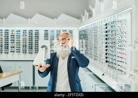 Contenu élégant homme senior regardant dans le miroir tout en essayant et choisir des lunettes dans la boutique optique moderne Banque D'Images