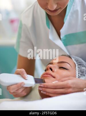 Gros plan des mains de l'esthéticienne faisant le traitement de la cavitation sur le visage humain avec l'équipement, la femme est couché et sourire doucement, le nettoyage et remo Banque D'Images