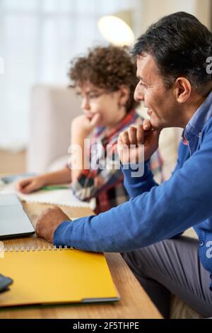 Préparer l'enfant au succès. Père d'âge moyen latin vérifiant ses devoirs, aidant son fils mignon, garçon d'école avec des études tout en étant assis à la table à Banque D'Images