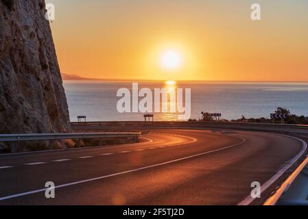 Route de montagne sans véhicules, en courbe et avec le soleil de l'aube à l'horizon au-dessus de la mer Méditerranée. Banque D'Images