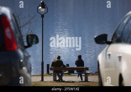 28 mars 2021, Bavière, Füssen: Les excursions s'assoient sur les rives de l'Hopfensee, à une distance l'une de l'autre, sur un banc au soleil derrière les voitures garées. Photo : Karl-Josef Hildenbrand/dpa Banque D'Images