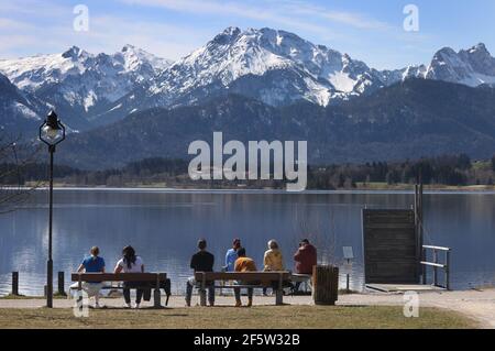 28 mars 2021, Bavière, Füssen: Des excursions s'assoient sur des bancs au soleil, sur les rives de l'Hopfensee, à une distance l'une de l'autre. Photo : Karl-Josef Hildenbrand/dpa Banque D'Images