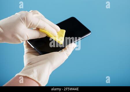 Nettoyage d'un smartphone avec une serviette jaune stérile dans des gants en caoutchouc sur fond bleu. Désinfection et prévention du virus Banque D'Images