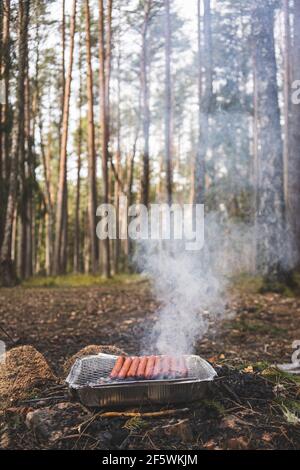 Griller des saucisses sur un gril instantané jetable. Griller des picniking dans la nature, entouré d'arbres forestiers et de pins Banque D'Images