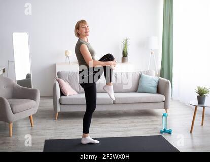 Heureuse femme senior faisant des exercices de flexibilité, étirant sa jambe sur le tapis de yoga à la maison, pleine longueur Banque D'Images