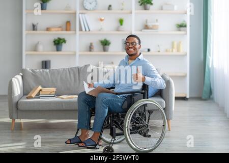 Homme noir gai handicapé en fauteuil roulant tenant un ordinateur portable, montrant le pouce vers le haut à la maison Banque D'Images