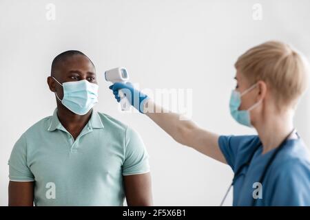 Femme d'âge moyen en masque de protection, gants et uniforme mesure la température avec un thermomètre infrarouge moderne Banque D'Images