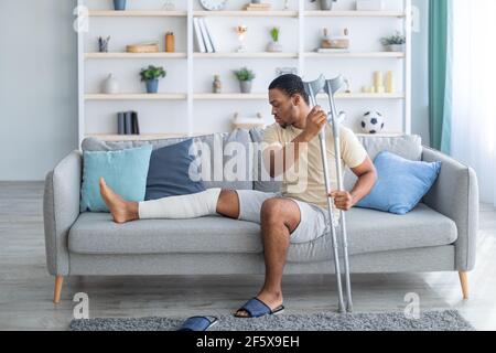 Jeune Afro-américain avec une jambe plâtrée assise sur un canapé, penchée sur des béquilles à la maison Banque D'Images