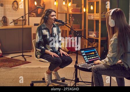 Jeune femme travaillant sur un ordinateur portable avec une jeune femme assise en face elle et chantant dans le microphone dans le studio d'enregistrement Banque D'Images