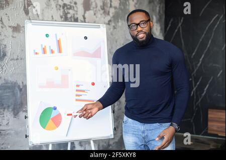 Un travailleur masculin qui présente une présentation à ses patrons, debout près d'un tableau blanc montrant des diagrammes, sur la formation d'entreprise, une réunion ou un séminaire, indique et explique le diagramme, donne un rapport sur les statistiques Banque D'Images