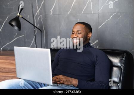 Spécialiste INFORMATIQUE afro-américain assis sur un grand fauteuil confortable, regardant l'écran d'un ordinateur portable. Joyeux homme noir lisant le message e-mail avec de bonnes nouvelles, discutant avec des clients en ligne, travaillant à distance Banque D'Images