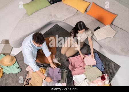 Jeune couple marié en vêtements décontractés assis sur le sol grand canapé dans la salle de séjour et emballage de leurs vêtements pendant le séjour pour avoir le voyage Banque D'Images