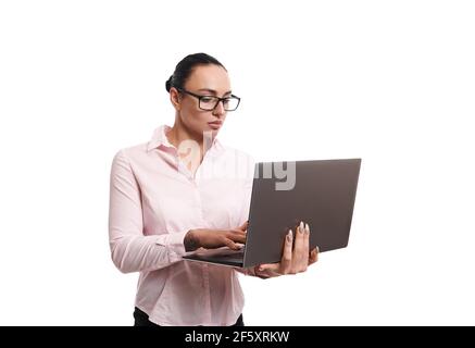Jeune femme d'affaires brunette portant des lunettes et une chemise rose dactylographiée texte sur l'ordinateur portable en se tenant isolé sur fond blanc avec copier l'espace pour Banque D'Images