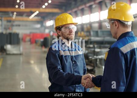 Deux hommes ingénieurs en casques de protection et en vêtements de travail se secouant les mains l'un contre l'autre dans un environnement d'atelier de grande envergure après avoir fait affaire Banque D'Images