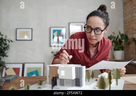 Jeune élégante femme architecte avec papier et stylo se pliant modèle de nouvelle maison et cour et pointant sur le toit tout en travaillant sur le projet Banque D'Images