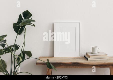 Maquettes blanches pour cadres de portrait sur banc, table vintage. Tasse de café sur pile de livres et plante en pot de monstère. Fond de mur blanc. Scandinave Banque D'Images