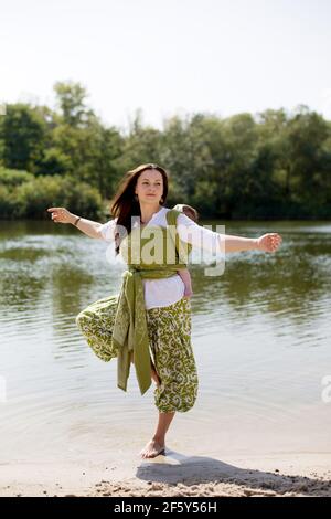 Mère active faisant du yoga à la rive de la rivière avec enveloppé dos petite fille Banque D'Images