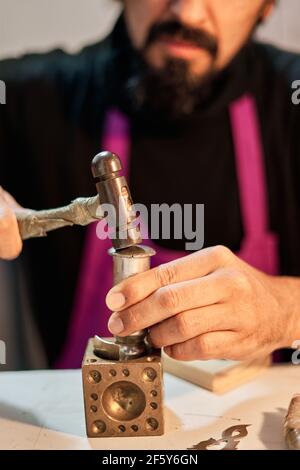 Photo verticale gros plan artisan bijoutier frapper une plaque de métal avec le marteau sur la table de travail Banque D'Images