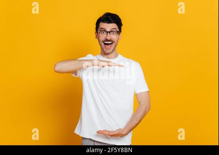 Stupéfait excité heureux caucasien homme en t-shirt blanc et des lunettes tient ses mains en face de lui, montre place pour votre publicité, se tenant sur un fond orange isolé, souriant et moqueuse Banque D'Images