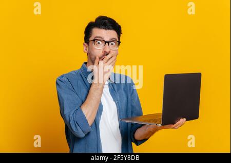 Choqué confus style non shaven caucasien avec des lunettes tenant un ordinateur portable ouvert dans sa main, regarde surpris à la caméra tout en se tenant contre un arrière-plan isolé orange Banque D'Images
