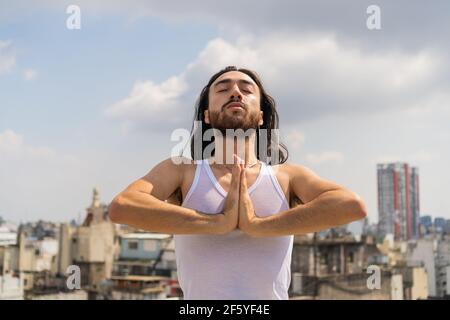 jeune homme faisant du yoga sur le toit urbain, avec la ville et le ciel à l'arrière-plan. homme avec les cheveux longs et la barbe portant un t-shirt sans manches. muscle Banque D'Images