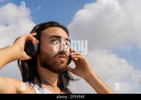 jeune homme barbu portant un casque sans fil avec le ciel en arrière-plan, réfléchissant sur la vie, méditant Banque D'Images