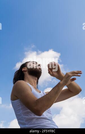 jeune homme méditant sur un toit. photo verticale d'un homme aux cheveux longs, mains dans l'air avec fond bleu ciel avec des nuages, photo artistique. usure Banque D'Images
