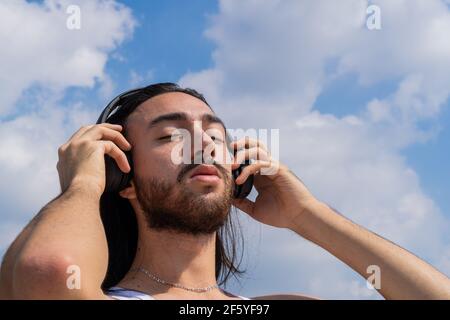 jeune homme barbu avec casque sans fil avec le ciel en arrière-plan, réfléchissant sur la vie, méditant. avec les yeux fermés. son surround. Banque D'Images