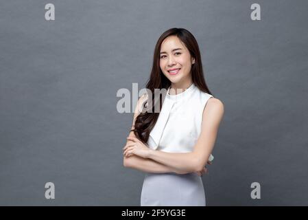 Souriante jeune femme d'affaires asiatique belle regardant l'appareil photo et faire mouvement croisé du bras sur fond gris studio isolé Banque D'Images