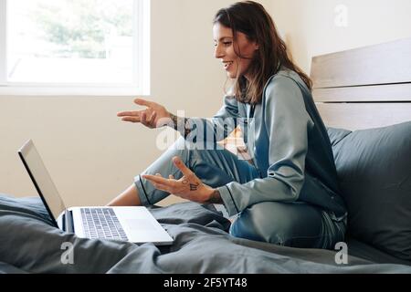 Belle jeune femme en pyjama de soie assis sur le lit et avoir une visioconférence avec des collègues Banque D'Images