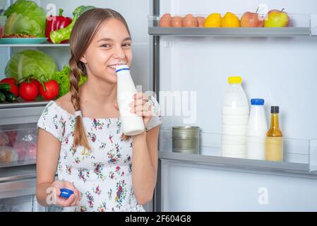 Belle jeune fille de l'adolescence tenant une bouteille de lait et des boissons tout en vous tenant près du réfrigérateur ouvert dans la cuisine à la maison Banque D'Images