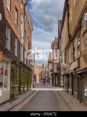 Une ancienne rue de York. Les vieux bâtiments sont de chaque côté et la lumière du soleil s'étend sur une jonction. Des boutiques bordent la rue et un ciel nuageux est au-dessus. Banque D'Images