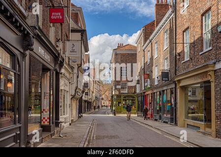 Une ancienne rue de York. Les vieux bâtiments sont de chaque côté et la lumière du soleil s'étend sur une jonction. Des boutiques bordent la rue et un ciel nuageux est au-dessus. Banque D'Images