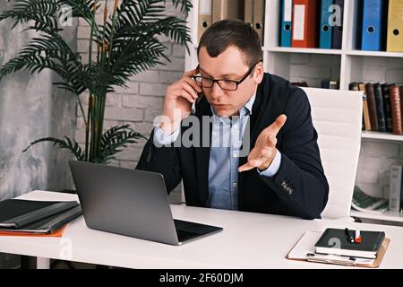 Portrait d'un homme indigné dans un costume classique et des lunettes regardant l'écran d'ordinateur portable criant dans le téléphone. Il est assis dans le bureau contre l'arrière-groupe Banque D'Images