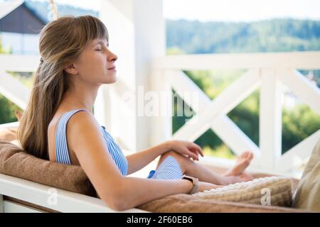 Jeune femme brune heureuse assise sur un canapé-terrasse avec des coussins moelleux profitant de la chaude journée d'été. Concept de temps libre sur l'air frais. Banque D'Images