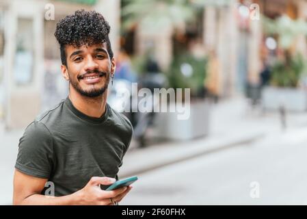 portrait d'un jeune et attrayant latino-américain regardant l'appareil photo et souriant. Il tient un téléphone cellulaire bleu. Il est dans la rue de Barcelone. Banque D'Images