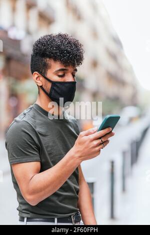 portrait vertical d'un jeune latino-américain qui vérifie son smartphone dans la rue. Il porte des vêtements d'été décontractés et un masque de protection noir. Banque D'Images