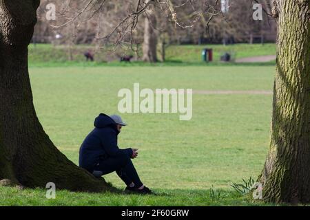 Londres, Royaume-Uni, 29 mars 2021: Sur Tooting Commons un homme est assis sur les racines d'un arbre et regarde son téléphone. Anna Watson/Alay Live News Banque D'Images