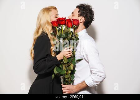 Charmant romantique jeune heureux couple célébrant un événement ensemble, femme tenant un bouquet de roses rouges isolé sur fond blanc, embrassant Banque D'Images