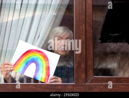 Une femme âgée dans la fenêtre de la maison tient le dessin d'un arc-en-ciel d'un enfant entre ses mains. Arc-en-ciel dans la fenêtre.Merci NHS, promotion en soutien Banque D'Images