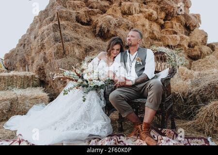 Un homme et une femme élégamment habillés dans une belle robe blanche s'assoient sur des chaises en osier sur le fond de balles de foin, style boho Banque D'Images