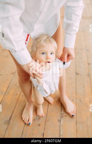 La jeune mère enseigne à marcher et conduit son petit fils par les mains sur un plancher en bois. Banque D'Images