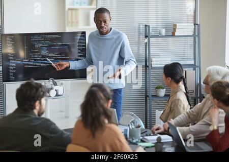 Portrait de l'homme afro-américain émotionnel donnant la présentation au bureau à l'équipe INFORMATIQUE et pointant sur le code à l'écran, l'espace de copie Banque D'Images