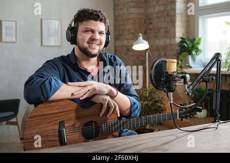 Portrait d'un jeune musicien barbu dans un casque assis avec de la guitare et sourire à l'appareil photo en studio Banque D'Images