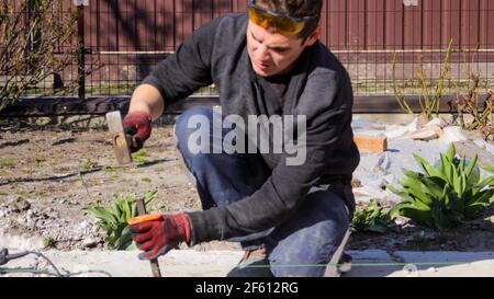 Recentrer un jeune ouvrier de construction avec un front ridé dans des lunettes jaunes éliminant les irrégularités sur la table de plancher avec un marteau et un burin. Mains de sexe masculin Banque D'Images