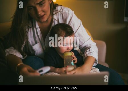 Mère vérifiant la température de bébé fille tout en prenant des conseils à travers consultation en ligne à domicile pendant une pandémie Banque D'Images